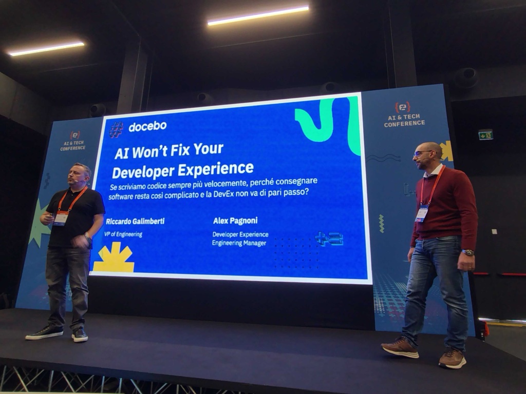 Two presenters stand on a stage beside a large blue slide that reads 'AI Won't Fix Your Developer Experience' at a tech conference.